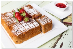 Square cake with cut slice on white plate with strawberries on top and couli on the side
